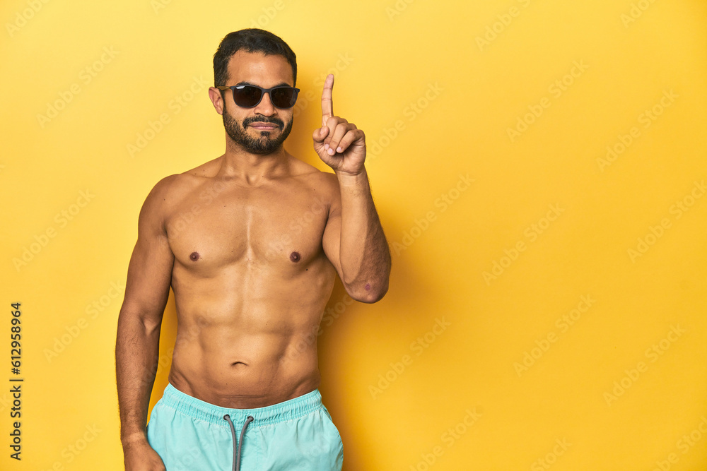 Fit young Latino man in swimwear and sunglasses, yellow studio background, showing number one with finger. Fit young Latino man in swimwear and sunglasses, yellow studio background, showing number one with finger.