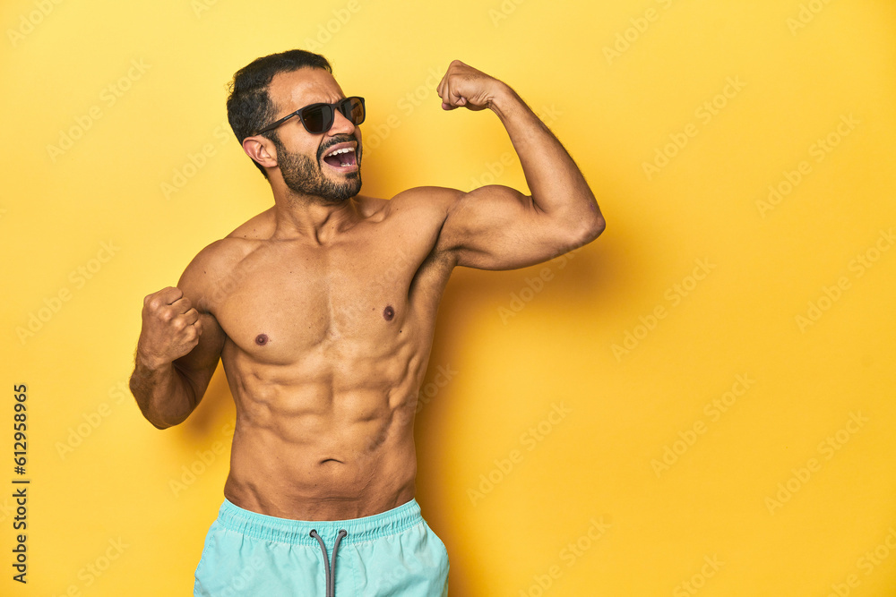 Fit young Latino man in swimwear and sunglasses, yellow studio background, raising fist after a victory, winner concept.