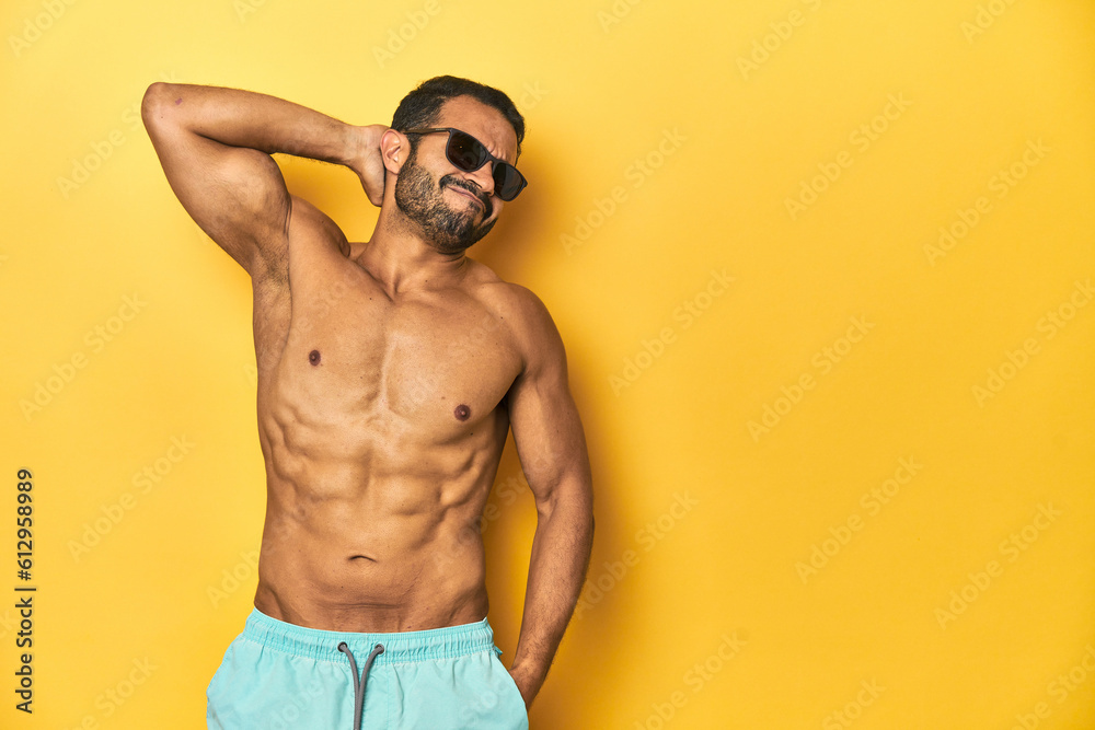 Fit young Latino man in swimwear and sunglasses, yellow studio background, touching back of head, thinking and making a choice.
