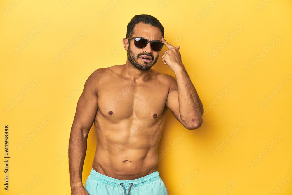 Fit young Latino man in swimwear and sunglasses, yellow studio background, showing a disappointment gesture with forefinger. Fit young Latino man in swimwear and sunglasses, yellow studio background, showing a disappointment gesture with forefinger.