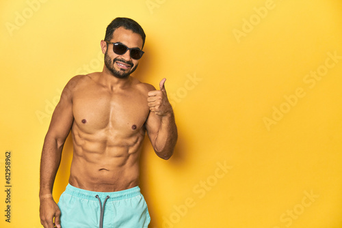 Fit young Latino man in swimwear and sunglasses, yellow studio background, smiling and raising thumb up
