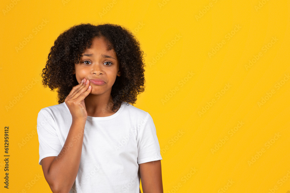 Sad curly teenager black schoolgirl in white t-shirt presses hand to cheek, suffering from toothache Sad curly teenager black schoolgirl in white t-shirt presses hand to cheek, suffering from toothache