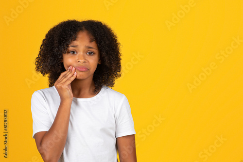 Sad curly teenager black schoolgirl in white t-shirt presses hand to cheek, suffering from toothache photo