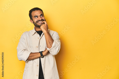 Casual young Latino man against a vibrant yellow studio background, relaxed thinking about something looking at a copy space.