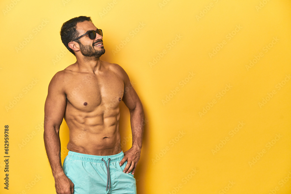 Cheerful muscular Latino man in swimsuit posing with ad space, yellow studio background.