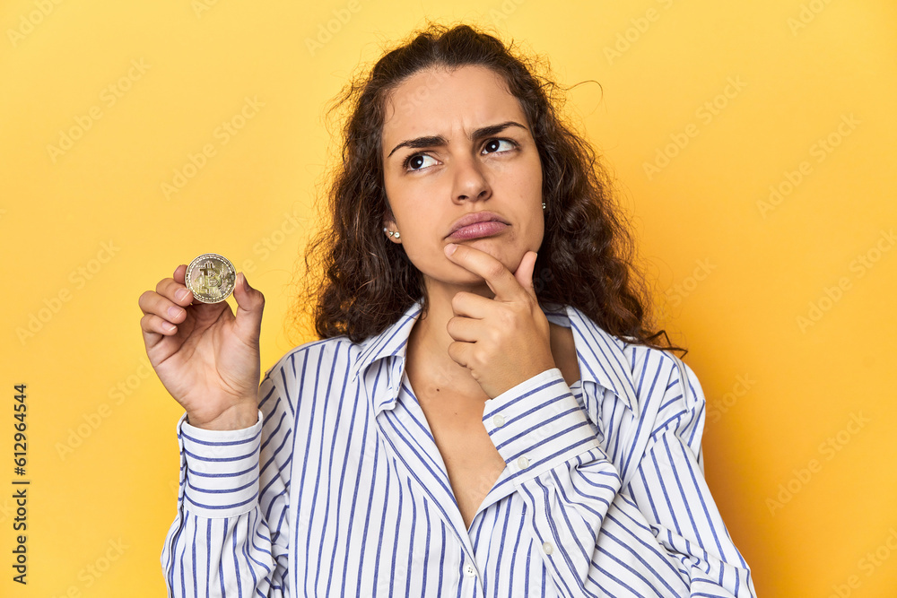 Woman holding a physical Bitcoin coin, looking sideways with doubtful and skeptical expression. Woman holding a physical Bitcoin coin, looking sideways with doubtful and skeptical expression.
