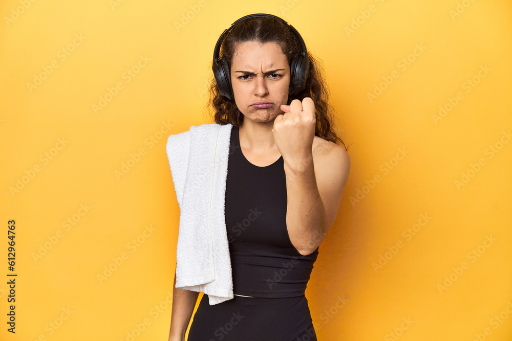 Athletic woman holding a towel, yellow background, showing fist to camera, aggressive facial expression. Athletic woman holding a towel, yellow background, showing fist to camera, aggressive facial expression.