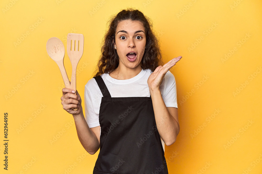 Woman with apron, wooden cooking utensils, yellow, surprised and shocked. Woman with apron, wooden cooking utensils, yellow, surprised and shocked.