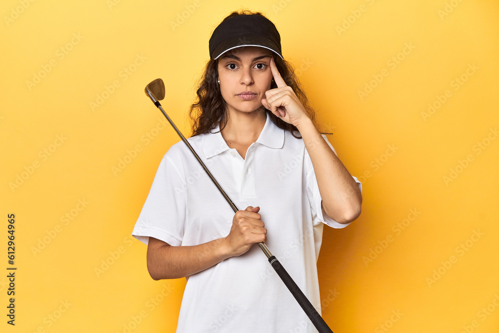 Golfer woman with cap, golf polo, yellow studio, pointing temple with finger, thinking, focused on a task. Golfer woman with cap, golf polo, yellow studio, pointing temple with finger, thinking, focused on a task.