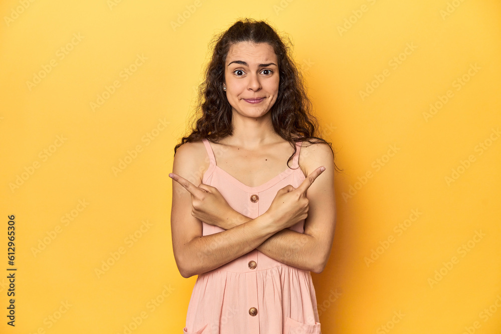 Young Caucasian woman, yellow studio background, points sideways, is trying to choose between two options.