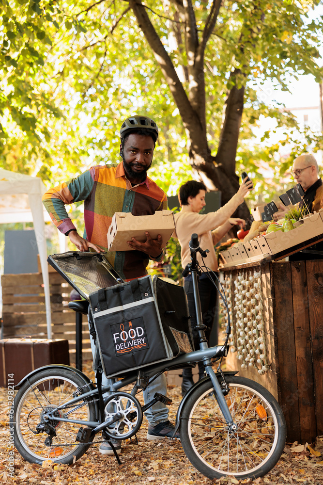 Organic locally grown fruits and vegetables delivery at farmers market, courier putting bio organic produce in thermal backpack and picking up order. Delivering food order fron local natural stand. Organic locally grown fruits and vegetables delivery at farmers market, courier putting bio organic produce in thermal backpack and picking up order. Delivering food order fron local natural stand.