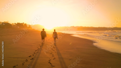 SILHOUETTE: Surf couple walking along gorgeous sandy beach towards golden sunset
