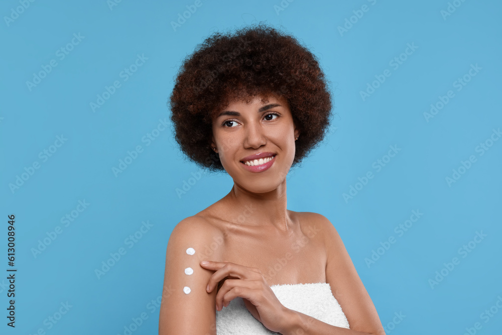 Beautiful young woman applying body cream onto arm on light blue background