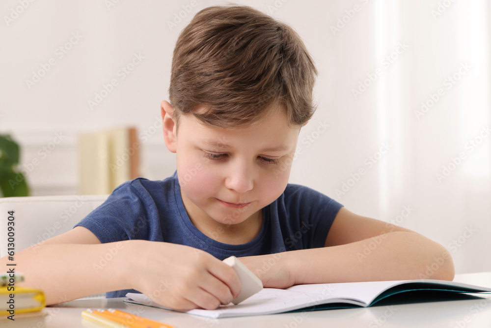 Little boy erasing mistake in his notebook at white desk indoors Little boy erasing mistake in his notebook at white desk indoors