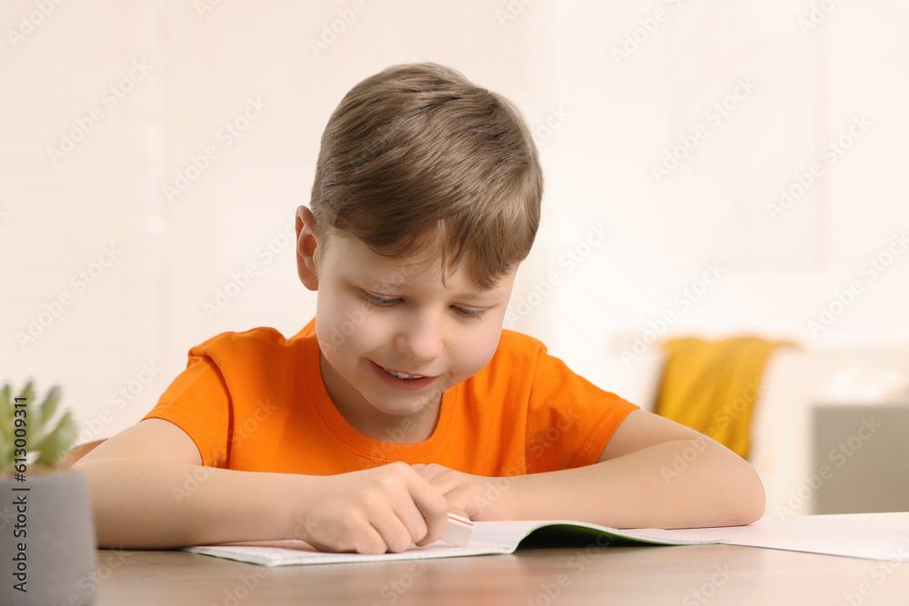 Little boy erasing mistake in his notebook at wooden desk indoors Little boy erasing mistake in his notebook at wooden desk indoors