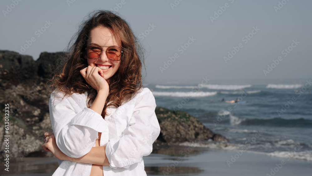 Pretty girl in white shirt and glasses having fun on the beach. Smiling at the camera, looking into the distance. Background of the ocean, waves, sky. A journey through Asia. Vacation in Bali.