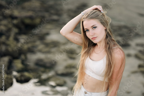 A young blonde woman in denim shorts and a white tank top enjoys the beauty of Bali. Portrait of a cute girl. Ocean coast. Sunset lighting. Tourism on the tropical beaches of Asia.