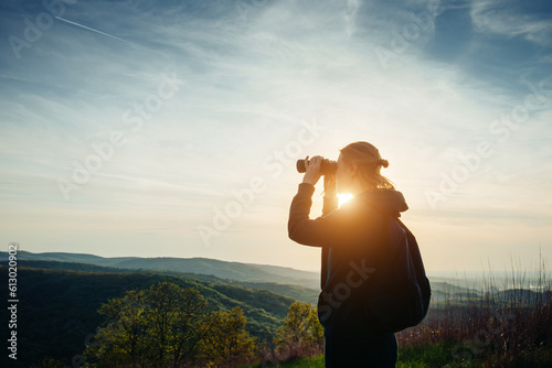 A hiker girl looks through binoculars at nature and birds standing on the top of a mountain in the bright rays of setting sun against the background of beautiful spring nature and cloudy blue sky