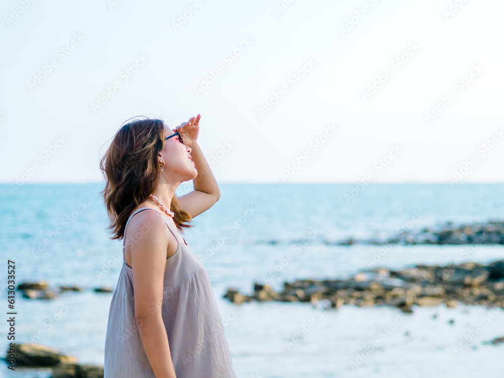 Beautiful Asian woman with sunglasses on the beach in summer vibes. Happy smile female covering the sunshine with her hand on the sea, rocks coast, island and sky background, Holiday vacation. Beautiful Asian woman with sunglasses on the beach in summer vibes. Happy smile female covering the sunshine with her hand on the sea, rocks coast, island and sky background, Holiday vacation.