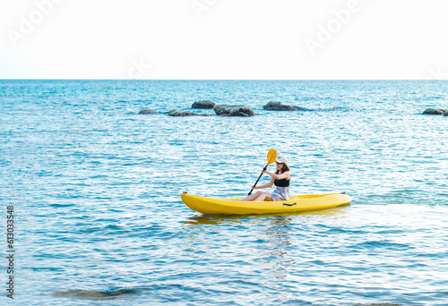 Happy Asian woman in black tank top and cap kayaking on yellow kayak boat with using paddle on the sea. Happy female having fun activity on seascape view background, Holiday trip vacation.
