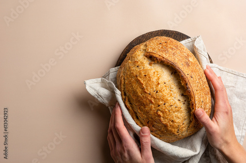 The baker's hands hold a loaf of fresh sourdough wheat bread, top view. Copy space banner of rustic cereal bread in a linen towel in female hands on a wooden board on a beige background.