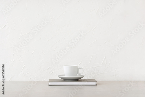 White cup with drink and notebook on neutral beige table, white concrete wall background with empty copy space. Minimalist brand template, coffee break, business lunch concept, home office workspace
