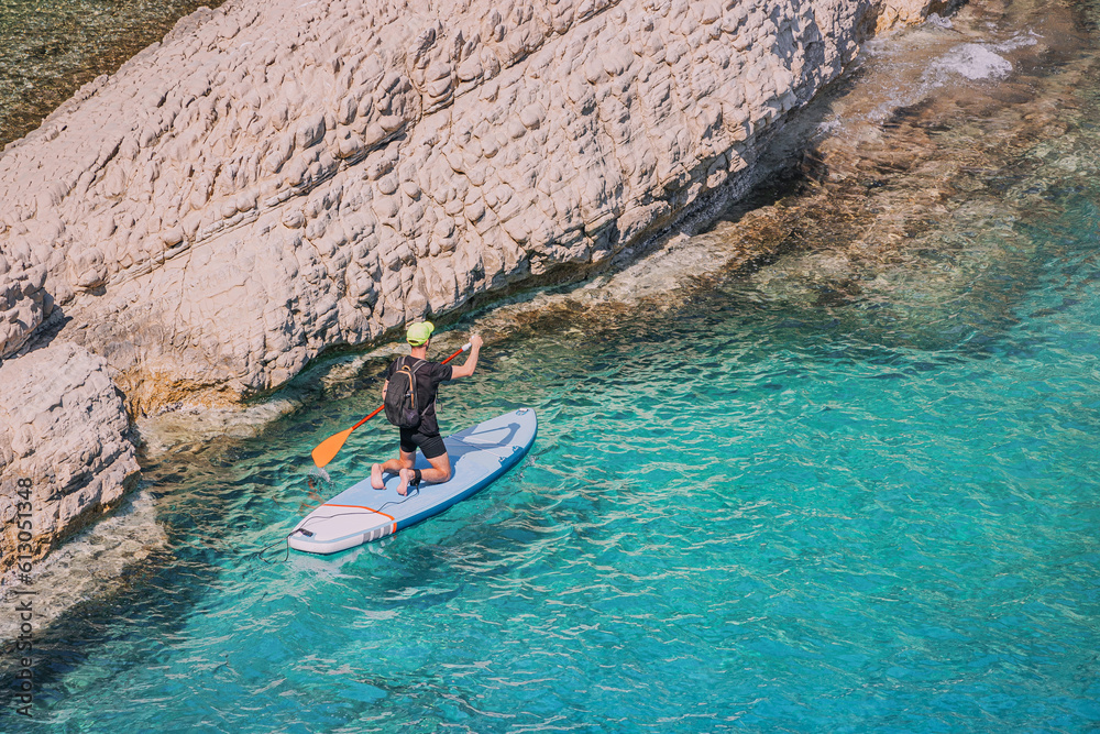 Fototapeta premium Happy man in a swimsuit with a paddle swims on a SUP board on the sea near huge rocks. Healthy lifestyle and recreation concept
