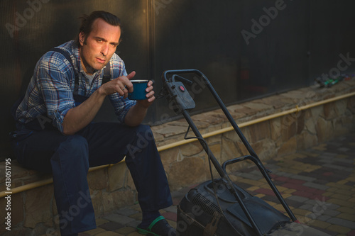 Attractive gardener man in blue work jumpsuit, looking at camera, sitting near a lawn mower with cup of coffee in hands, relaxing after hard working day in the garden. Sunset sun rays fall on his face