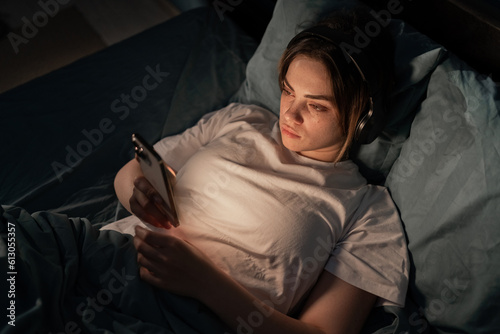 Young tired woman in headphones on bedtime listening music and chatting with friends, suffering from insomnia. Chat night and surfing phone. Social networks and phone addiction.