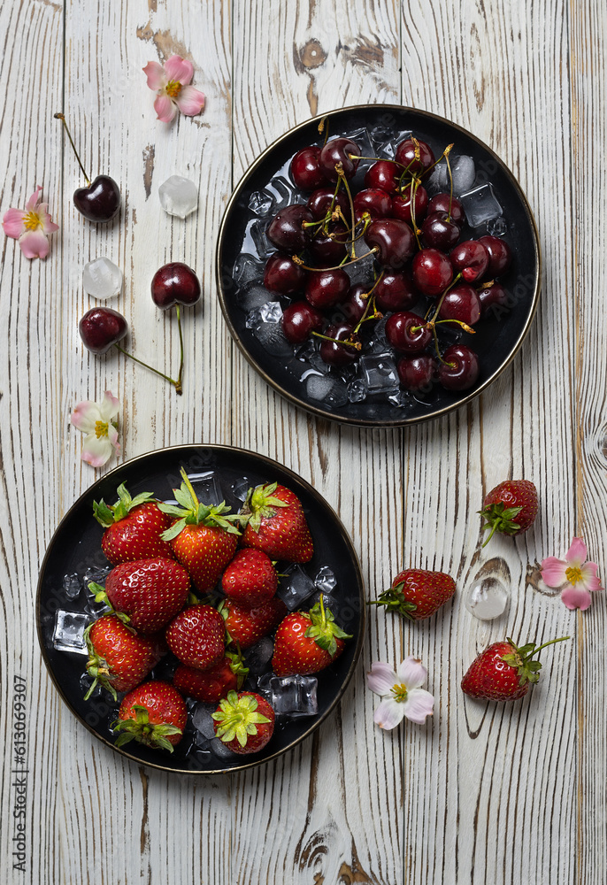 Ripe strawberries and cherries on black plates with ice. Ice cubes and pink flowers on the table. Light wooden background. Overhead view Ripe strawberries and cherries on black plates with ice. Ice cubes and pink flowers on the table. Light wooden background. Overhead view