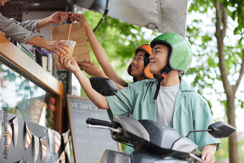 Happy young Asian couple in protective helmets taking paperbag with fast food and glass of soda while moving on bike by street truck