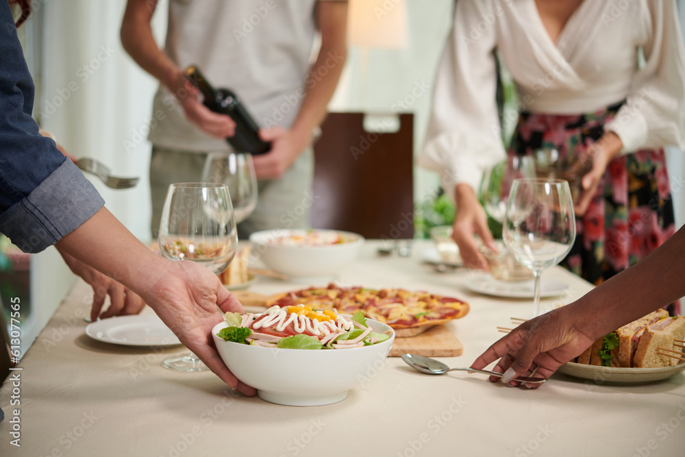 Close-up of young friends putting bowls of appetizing homemade salad and other snacks on table while serving it for dinner or home party