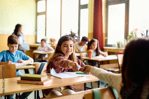 Happy schoolgirl gives pencil to her classmate during class in classroom.