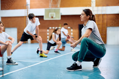 Female coach having class with elementary students at school gymnasium.