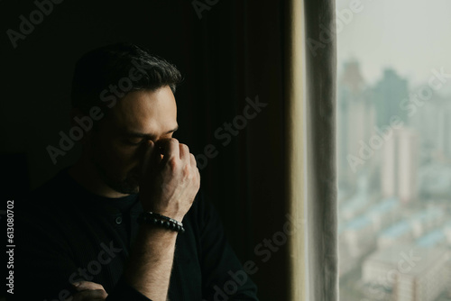 Frustrated man stands by a window and puts his hand to his face