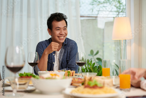 Young cheerful Asian man in casualwear sitting by served table at dinner prepared for guests and looking at his friend during conversation