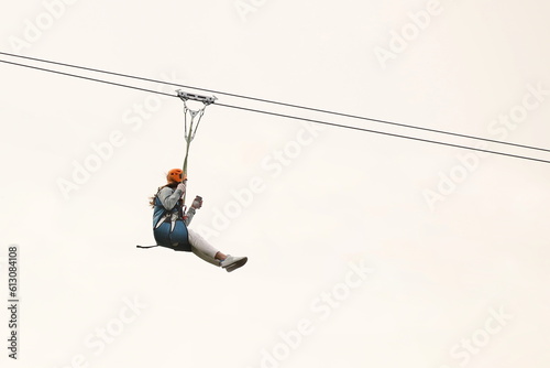 A woman in a rock climbing outfit slides down a steel cable on a roller gurney against the sky. Extreme recreation and sports. Amusement park for brave people. Outdoor activity. dangerous hobby