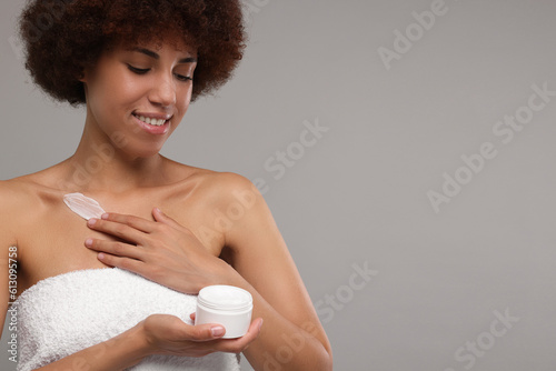 Beautiful young woman applying cream onto body on grey background, space for text