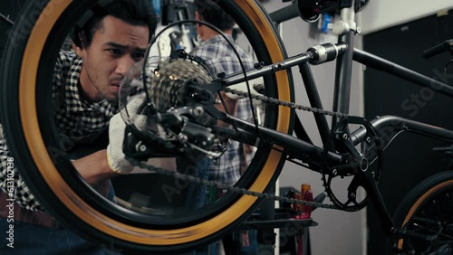 Proficient young man fixing and spinning wheel and gear of bike in cycle store. Young repairman studying details repairing bicycle in  bicycle shop. Maintenance and repair Concept