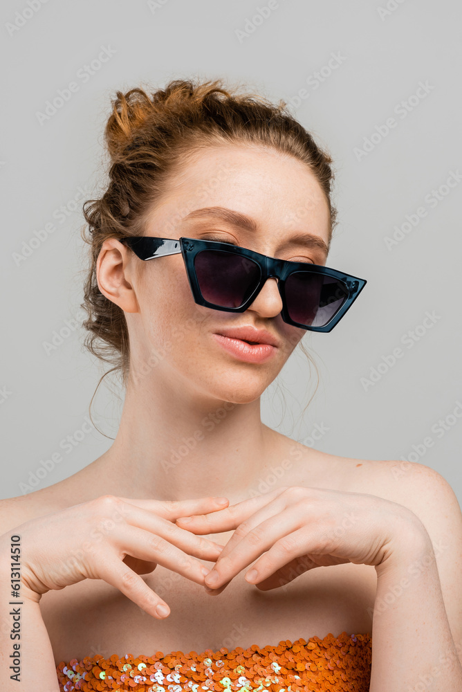 Portrait of young redhead woman with freckles wearing top with sequins and stylish sunglasses while standing isolated on grey background, trendy sun protection concept, fashion model Portrait of young redhead woman with freckles wearing top with sequins and stylish sunglasses while standing isolated on grey background, trendy sun protection concept, fashion model