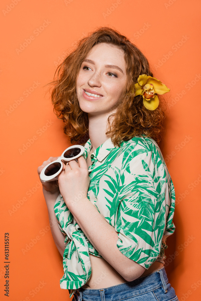 Smiling young redhead woman with orchid flower in hair wearing blouse with floral print and holding stylish sunglasses on orange background, summer casual and fashion concept, Youth Culture Smiling young redhead woman with orchid flower in hair wearing blouse with floral print and holding stylish sunglasses on orange background, summer casual and fashion concept, Youth Culture