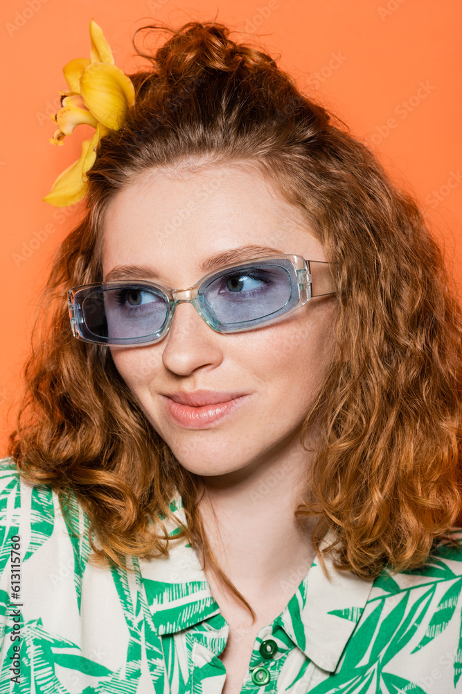 Portrait of young redhead model with orchid flower in hair wearing blue sunglasses and trendy blouse with floral pattern isolated on orange, stylish casual outfit and summer vibes concept Portrait of young redhead model with orchid flower in hair wearing blue sunglasses and trendy blouse with floral pattern isolated on orange, stylish casual outfit and summer vibes concept