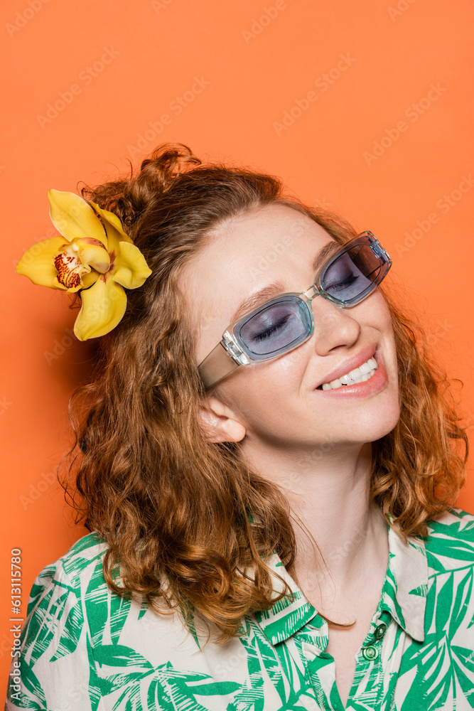 Smiling and trendy young woman with red hair and orchid flower posing in sunglasses and blouse with floral print on orange background, summer casual and fashion concept, Youth Culture Smiling and trendy young woman with red hair and orchid flower posing in sunglasses and blouse with floral print on orange background, summer casual and fashion concept, Youth Culture
