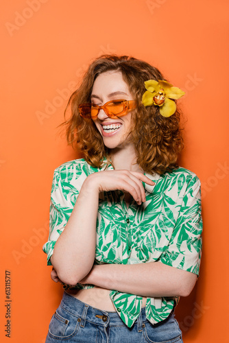 Happy young redhead woman with orchid flower and sunglasses posing in blouse with floral print and jeans on orange background, summer casual and fashion concept, Youth Culture