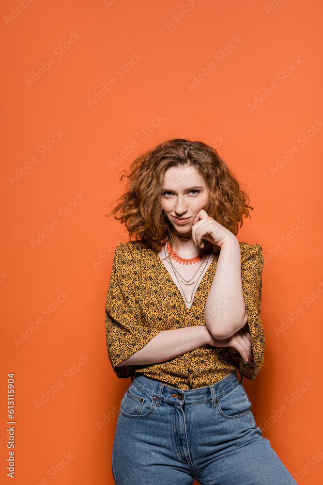 Confident and smiling red haired woman in blouse with abstract pattern and jeans looking at camera while standing on orange background, stylish casual outfit and summer vibes concept, Youth Culture Confident and smiling red haired woman in blouse with abstract pattern and jeans looking at camera while standing on orange background, stylish casual outfit and summer vibes concept, Youth Culture