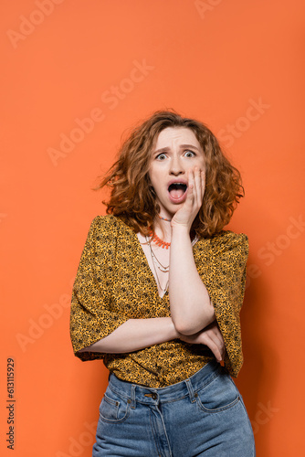 Shocked young redhead woman in blouse with abstract pattern and jeans looking at camera and standing on orange background, stylish casual outfit and summer vibes concept, Youth Culture © LIGHTFIELD STUDIOS