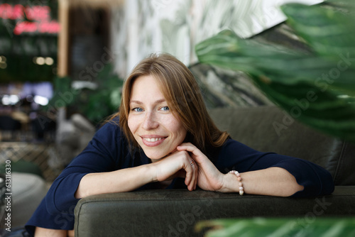 Happy young woman looking at camera. Portrait of comfortable woman relaxing on green armchair. Portrait of beautiful girl smiling and relaxing on green plants background