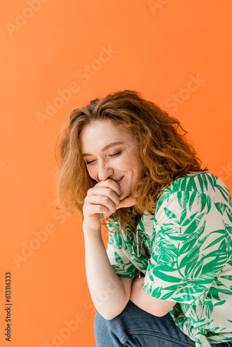 Cheerful young red haired woman with closed eyes in blouse with floral pattern and jeans laughing while posing isolated on orange, trendy casual summer outfit concept, Youth Culture
