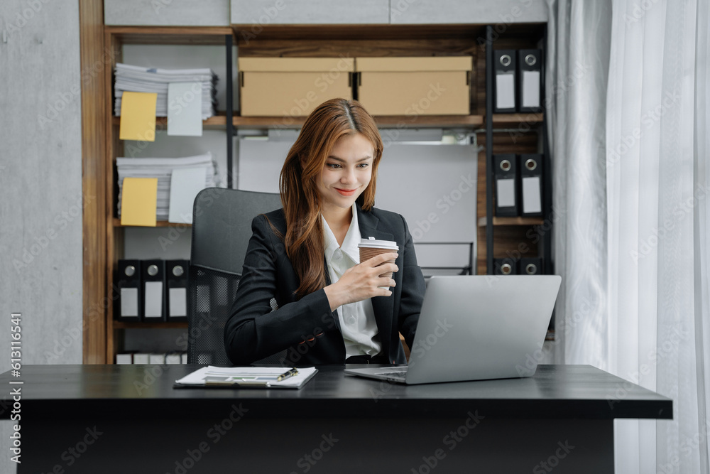 Asian businesswoman sits in a office working on laptop and enjoys a coffee.