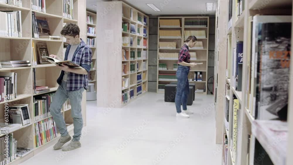 Two young youth persons are searching for a book in university library. They are between book shelves and look for a specific publication print. Young, well educated, smart and curious future leaders. Two young youth persons are searching for a book in university library. They are between book shelves and look for a specific publication print. Young, well educated, smart and curious future leaders.